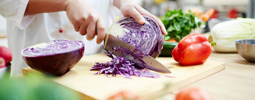 Hand holding red cabbage with the other hand slicing it on a chopping board